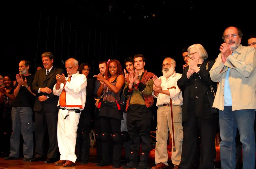 Elenco e convidados enfileirados no palco, aplaudindo ao final de uma apresentação