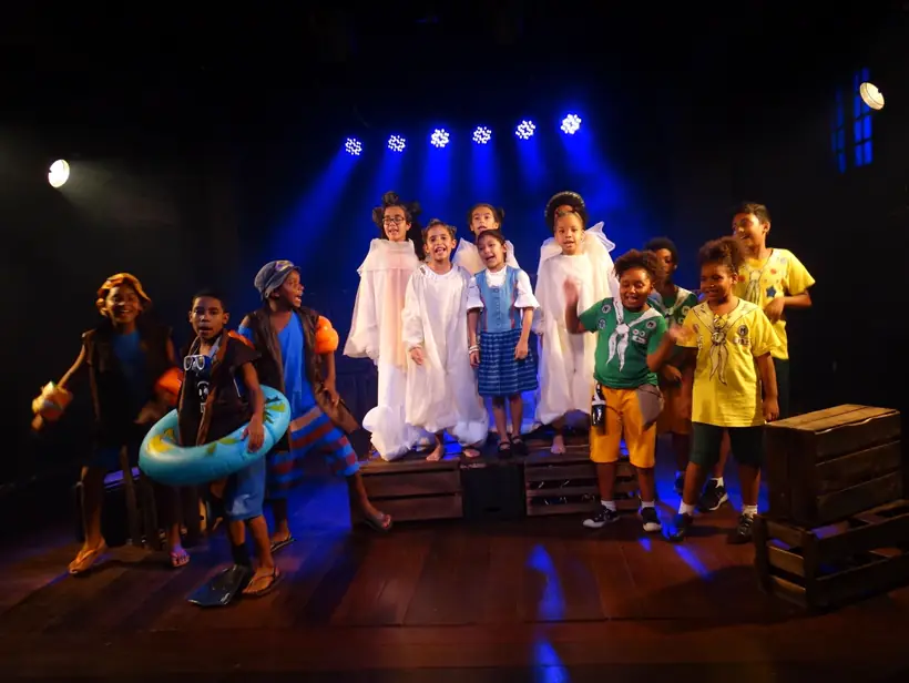 Crianças em cena sobre o palco, iluminadas por refletores azuis visíveis ao fundo