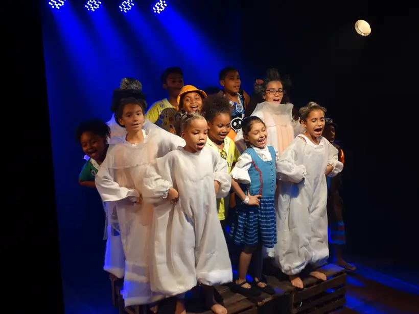 Crianças em camisolas brancas cantando sobre engradados, sob feixes de luz azul que descem da vara de refletores