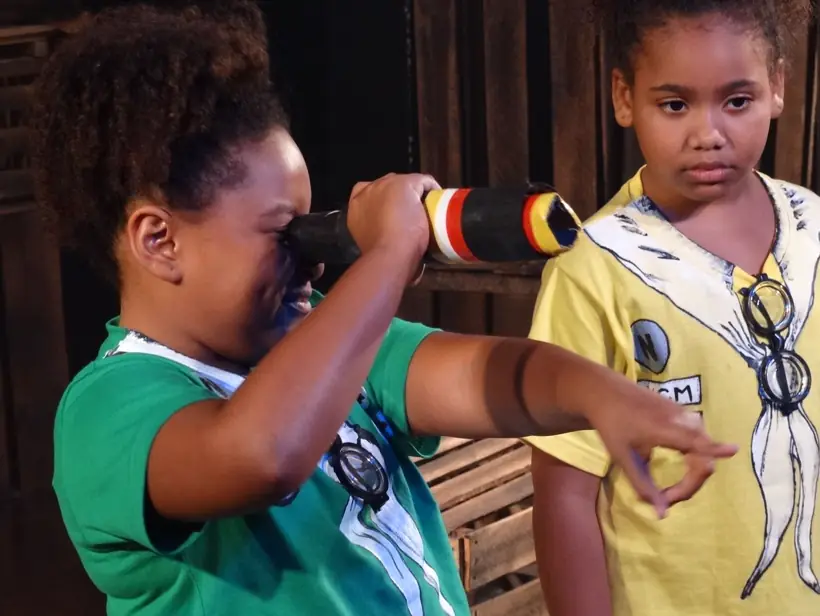 Menina olhando por um telescópio de brinquedo ao lado de outro ator, em cena