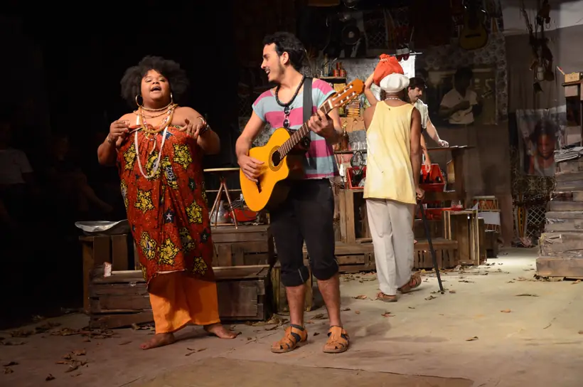Atores tocando violão e cantando em cenário de armazém, com elenco ao redor