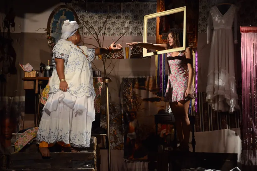 Atriz de vestido branco em cenário de sala com quadros pendurados na parede